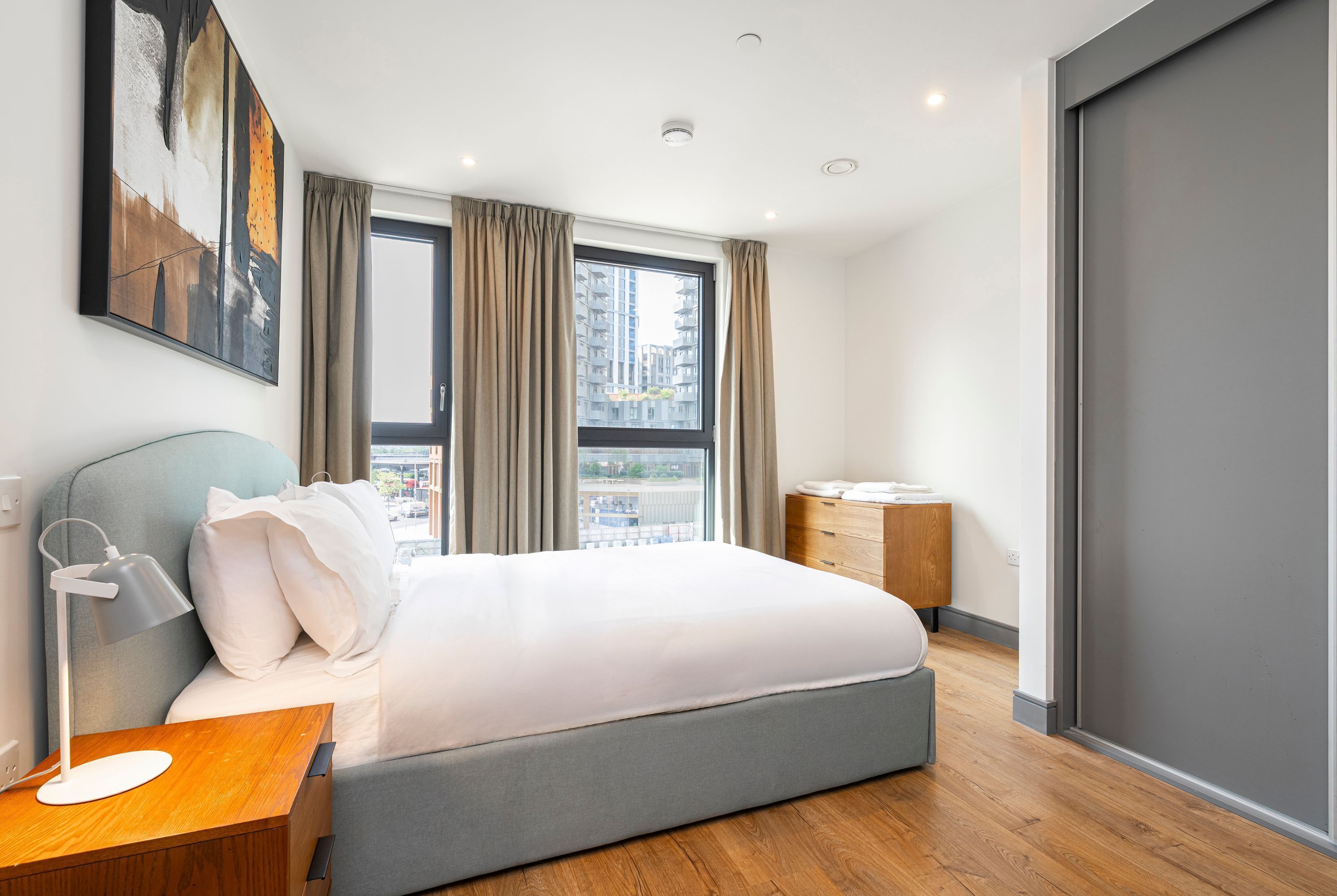 A modern hotel room with a large bed, a bedside table with a lamp, a dresser, and a view of the city through a large window.