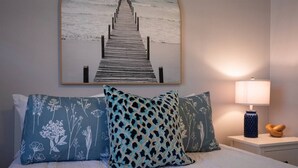 Un llit amb almoinades estampades florals, un amb motllures de fusta, una tauleta de nit amb una làmpada, i una obra d'art penjada a la paret que representa un moll.