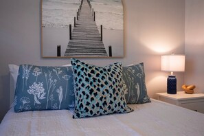 Ein Bett mit blumigen Kissen, ein hölzernes Kopfbrett, ein Nachttisch mit einer Lampe und ein an der Wand befestigtes Kunstwerk, das einen Steg zeigt.