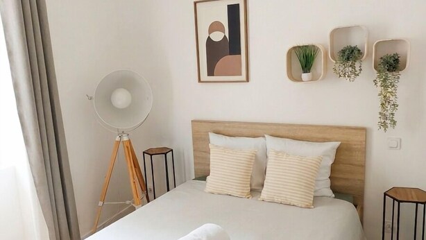 Una cama bien tendida con una toalla enrollada, un cabecero de madera, una obra de arte abstracta enmarco y tres macetas fijadas a la pared.