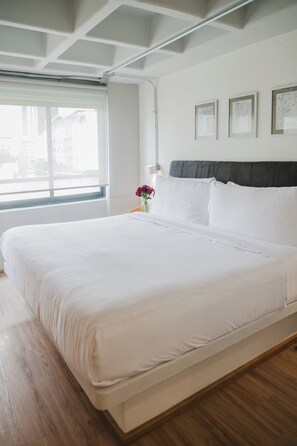 A large bed with white bedding, a wooden floor, a window with a view of buildings, and a wall-mounted light fixture. - Modern 1BR in Juárez with fast WiFi, great natural light, a comfortable king bed, a fully equipped kitchen, and a luxury bathroom. Perfect for couples or solo travelers seeking comfort, style, and a prime location in Mexico City. (Cuauhtémoc)