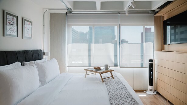 Ein modernes Hotelzimmer mit einem großen Bett, einem Nachttisch mit Tasse und Gebäck, einem an der Wand befestigten Fernseher und einem großen Fenster mit Blick auf Gebäude.