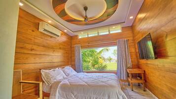 A bedroom with a bed, wooden walls, a ceiling fan, and a view of greenery outside.