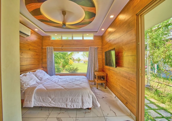 Une chambre à coucher avec un lit, des murs en bois, un ventilateur de plafond et une vue sur la verdure à l’extérieur.