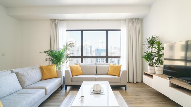 A modern living room with a sofa, coffee table, and TV, featuring a city view through large windows. - GMA Amwaj 2BR Sea View High Floor (Dubai)