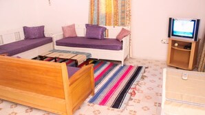 Una habitación con una cama de madera, un sofá cama con cojines morados, un televisor sobre un mueble de madera y una alfombra a rayas.