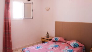 Un dormitorio con cama, mesita de noche, espejo adosado a la pared, ventana con cortinas y aplique de pared.