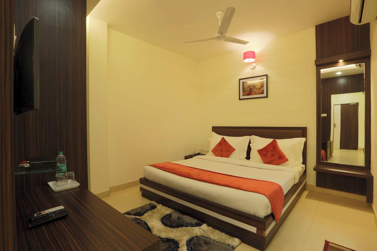 A hotel room with a bed, a ceiling fan, a mounted TV, a bedside table with a remote control, and a water bottle.