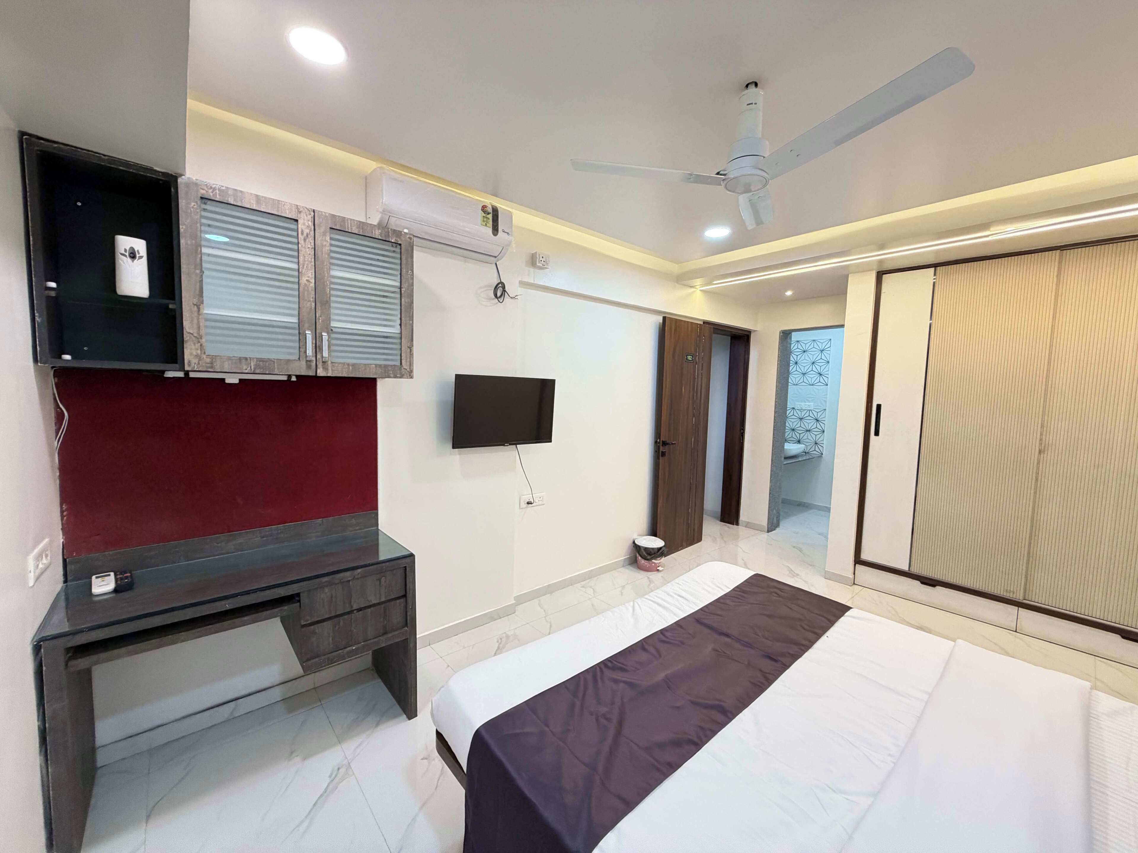 A bedroom with a bed, a desk, a TV, an air conditioner, a ceiling fan, and a wardrobe.