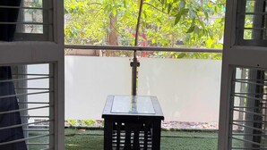 Vista a través de una ventana con una mesa y una silla en el exterior, rodeado de vegetación y un muro blanco.