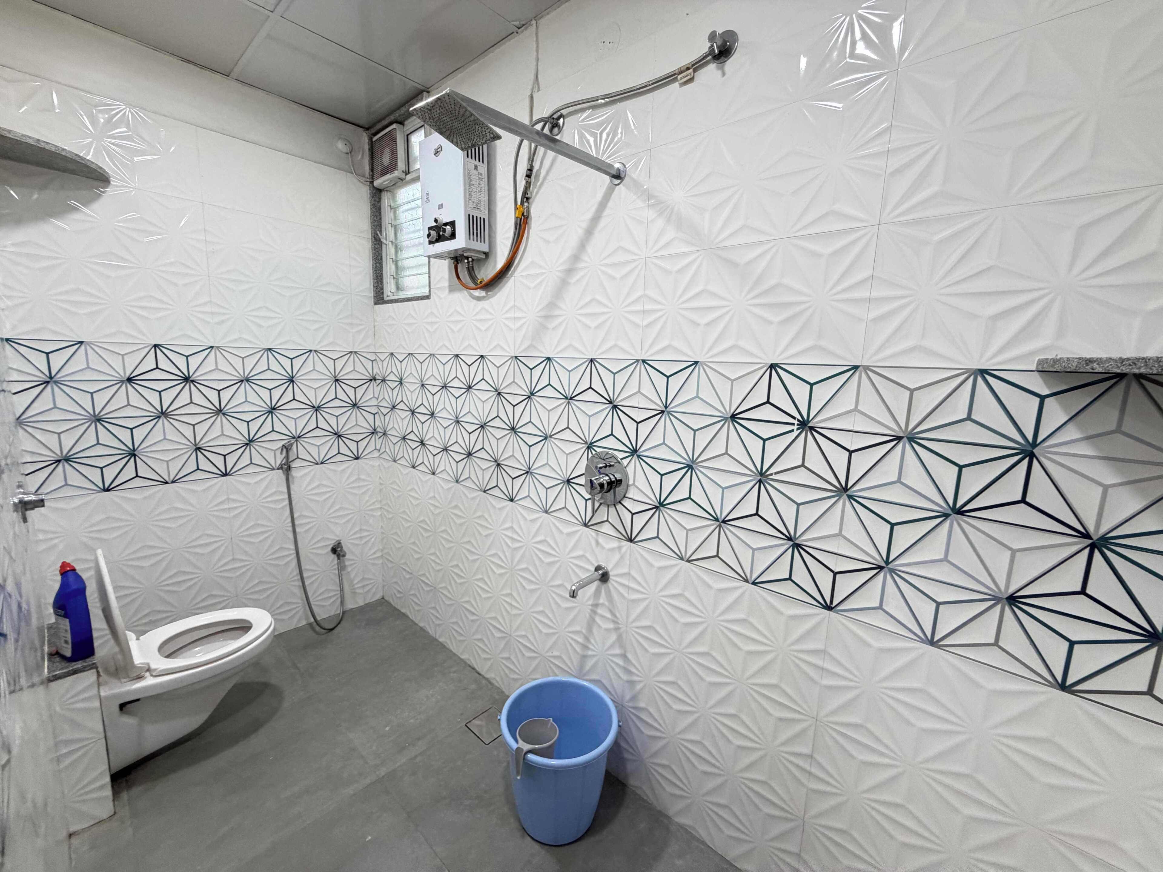 Un baño con inodoro blanco, un cubo azul y una ducha con un diseño geométrico de azulejos.
