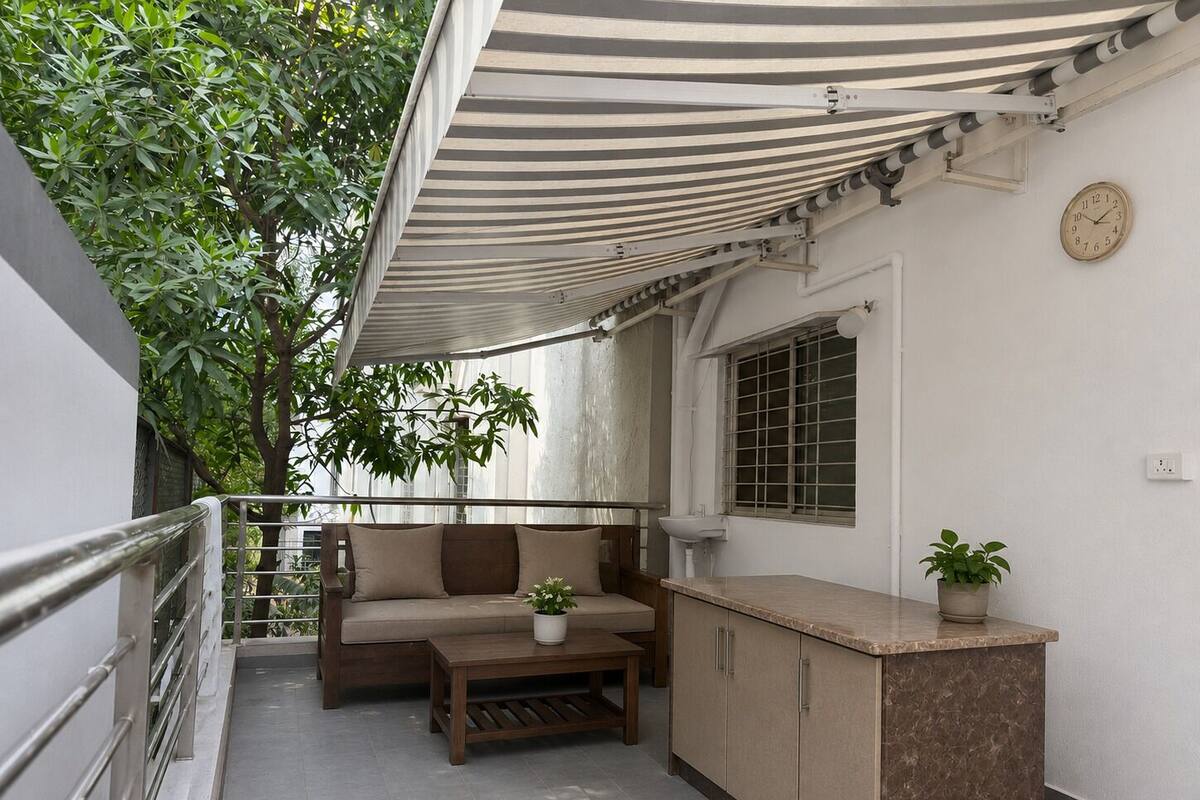 Een balkon met een gestreept zonnescherm, een bank, een bijzettafeltje, een bloempot, een gootsteen en een klok aan de muur.