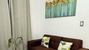 A brown sofa with two decorative pillows, a small round table with a book, a potted plant, and a painting on the wall. - Enjoy your getaway at this unique, private, quiet and safe residential house! (San Miguel)