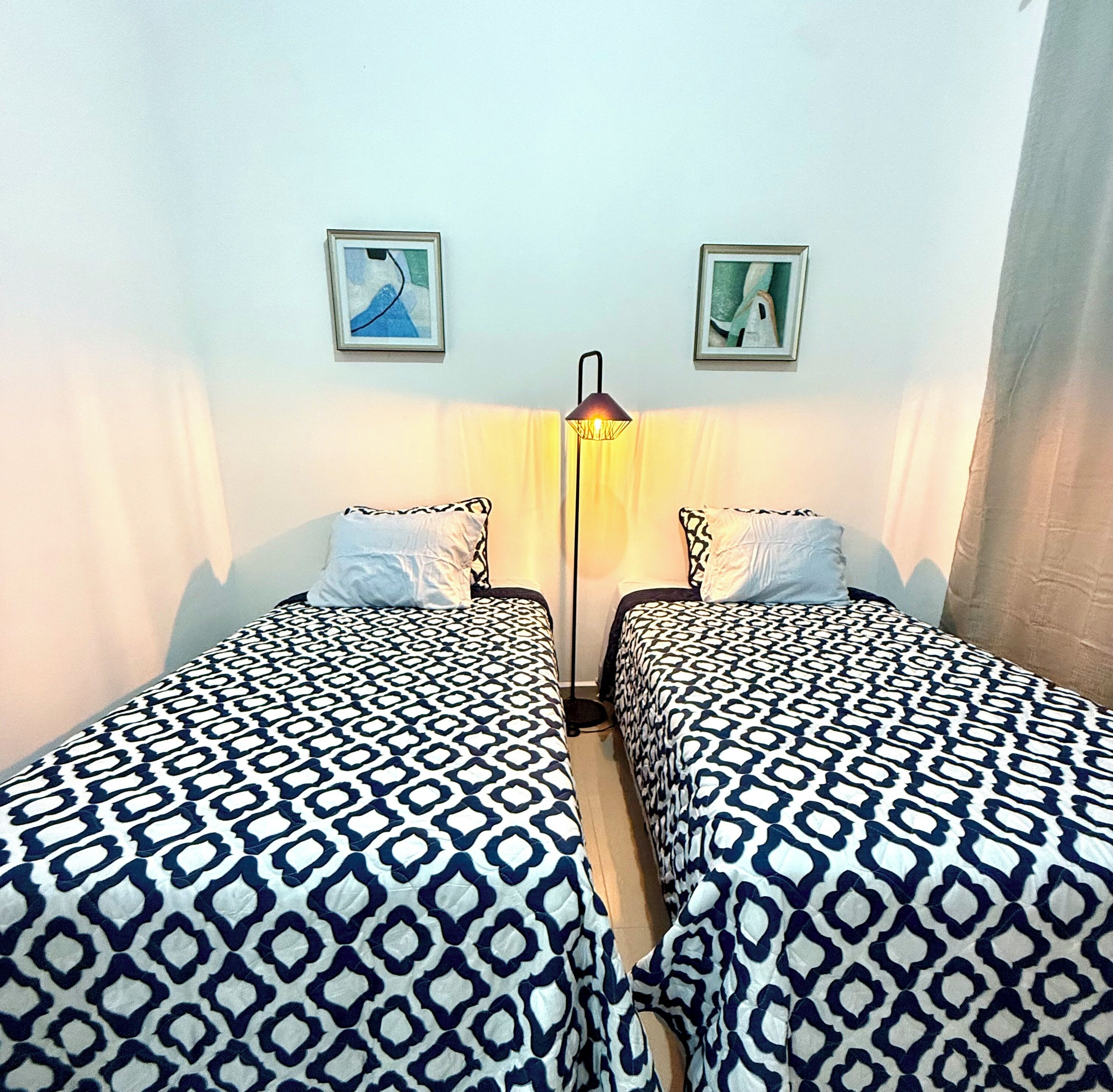 A room with two beds, each with patterned bedding, a floor lamp, and two framed pictures on the wall.
