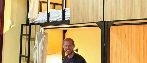 Habitación con literas, una persona sentada en la cama inferior, el número '2' en la pared y una cortina a la izquierda.