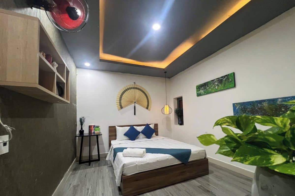 Une chambre à coucher comprenant un lit, une table de chevet, un ventilateur fixé au mur et une plante en pot.