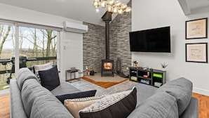 A modern living room with a fireplace, grey sofas, a flat-screen TV, and a large window overlooking a deck. - Ski in/out @ Camelback: Sleeps up to 16 (Tannersville)