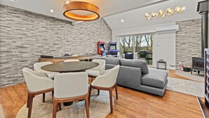 A modern living room with a gray sectional sofa, a round wooden dining table with white chairs, a built-in kitchen area with a stove, and a large window offering a view of trees. - Ski in/out @ Camelback: Sleeps up to 16 (Tannersville)