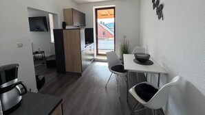 Eine kompakte Küche mit Backofen, ein Essbereich mit einem weißen Tisch und Stühlen sowie Blick auf ein rotes Gebäude durch eine Glastür.
