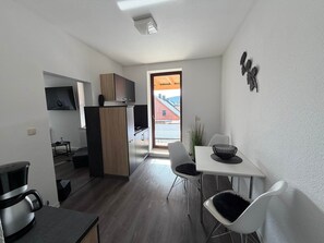 Una cocina compacta con horno empotrado, un comedor con mesa y sillas blancas, y vista a un edificio rojo a través de una puerta de vidrio.