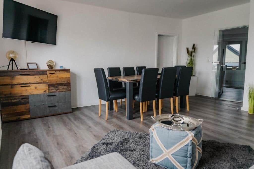 Ein modernes Wohnzimmer mit Esstisch, schwarzen Stühlen, einem Holzkabinett, einem Flachbildfernseher und einem grauen Teppich.