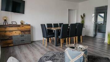 Ein modernes Wohnzimmer mit Esstisch, schwarzen Stühlen, einem Holzkabinett, einem Flachbildfernseher und einem grauen Teppich.
