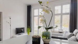 Een moderne woonkamer met een witte bank, een bijzettafeltje met een kaars en bloemen, en een televisie op een dressoir.