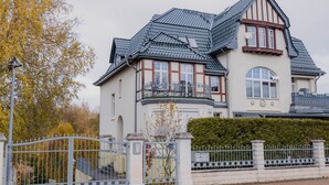 Een ruim, twee verdiepingen tellend huis met een opvallend balkon, een hekpoort en een verzorgde haag.