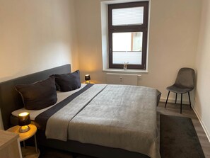 Un dormitorio con cama, lámparas de noche, una silla, una ventana y un radiador.