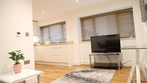 Ein modernes Wohnzimmer mit Flachbildfernseher, Glastisch, Blumentopf und einer Küche im Hintergrund.