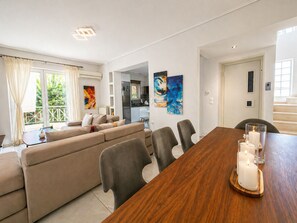 Uma sala de estar moderna com sofá, mesa de jantar e cadeiras, cozinha ao fundo e arte abstrata nas paredes.