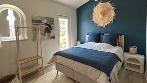 A bedroom with a bed, a wooden ladder, a painting, a woven light fixture, and a window.