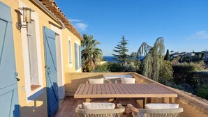 A wooden deck with a table and chairs, a blue door, and a view of the sea and hills.