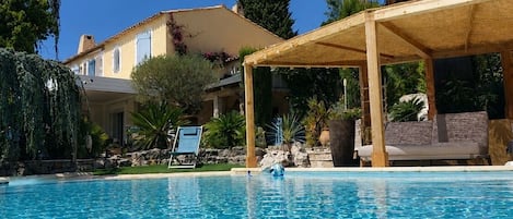 Une piscine dotée d’une terrasse en bois et de chaises longues, entourée de plantes tropicales et bordée d’un bâtiment au toit recouvert de tuiles.
