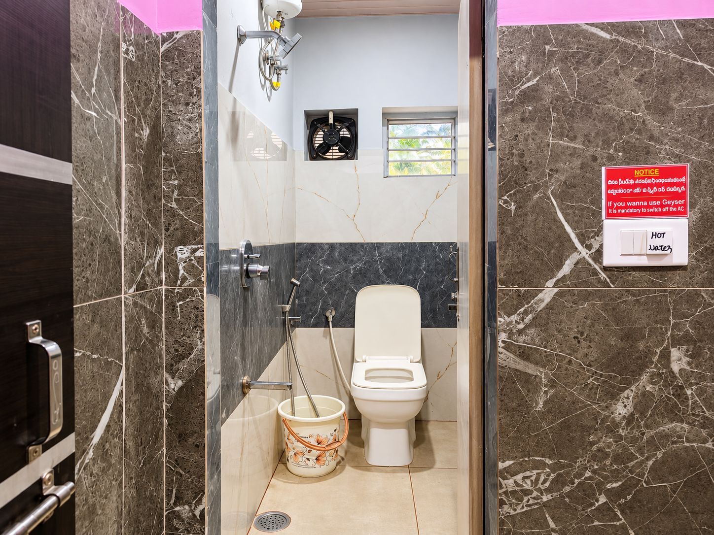 A bathroom with a glass-enclosed shower, a toilet, and a bucket. A notice is visible on the wall.