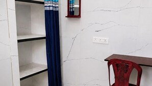 A bathroom with a white marble wall, a blue curtain, a red plastic chair, and a small mirror. - SVS Grands (Atreyapuram)