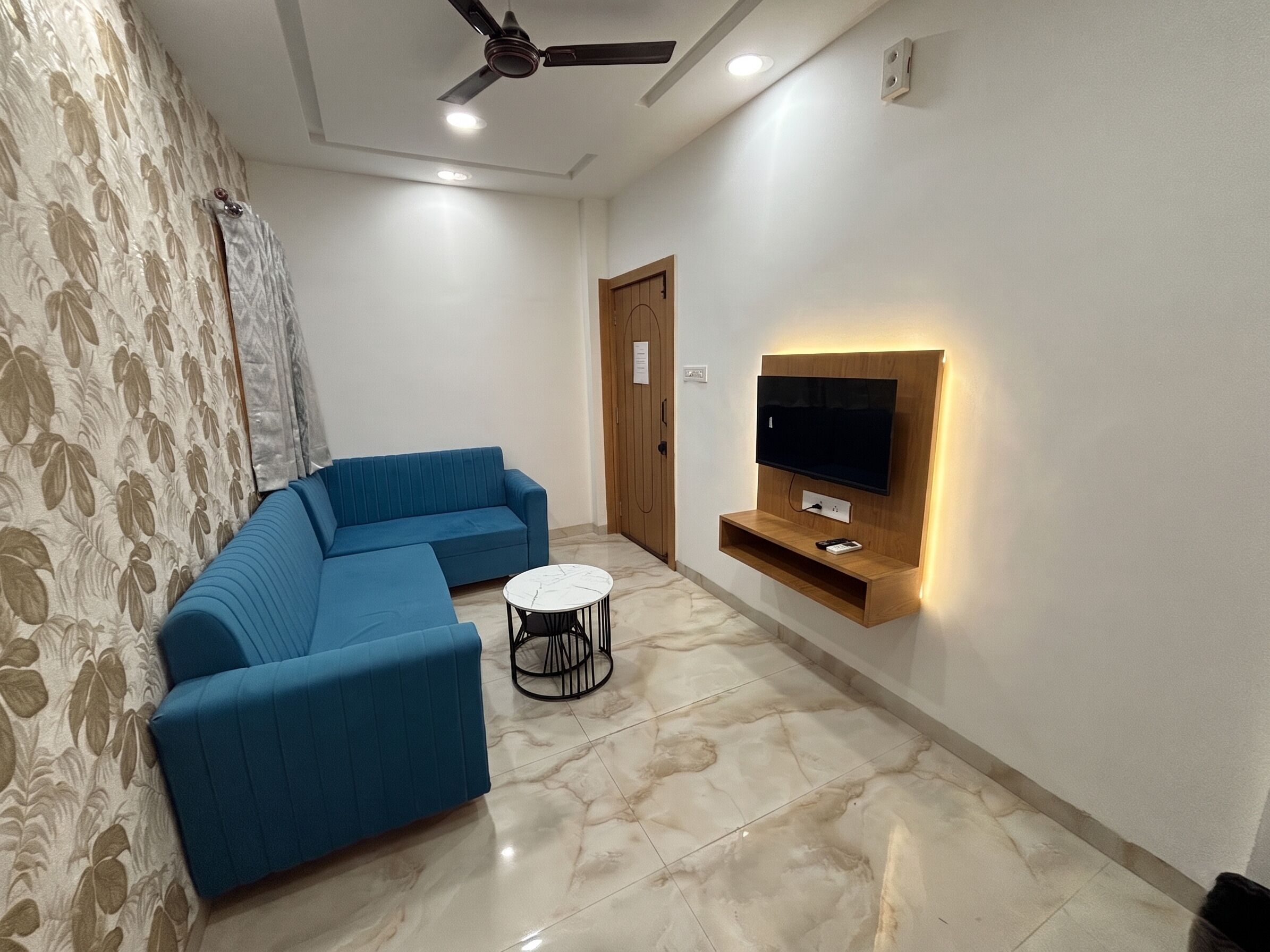 A living room with a blue sofa, a small round table, a ceiling fan, recessed lighting, a TV mounted on a wooden shelf, and a patterned curtain.