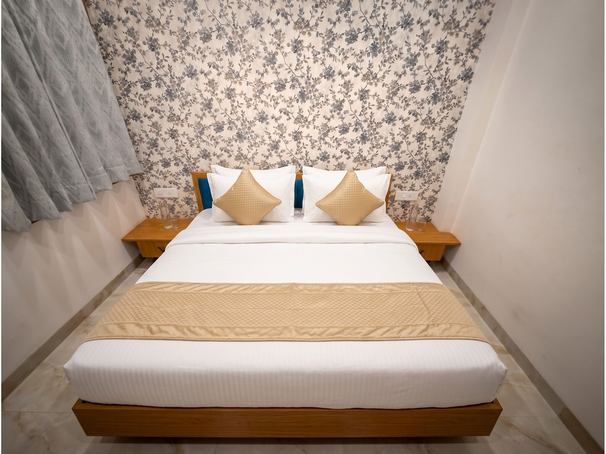 Un lit bien fait, recouvert d’une courtepointe matelassée, deux tables de chevet avec des lampes, un papier peint à motifs floraux et un rideau gris.
