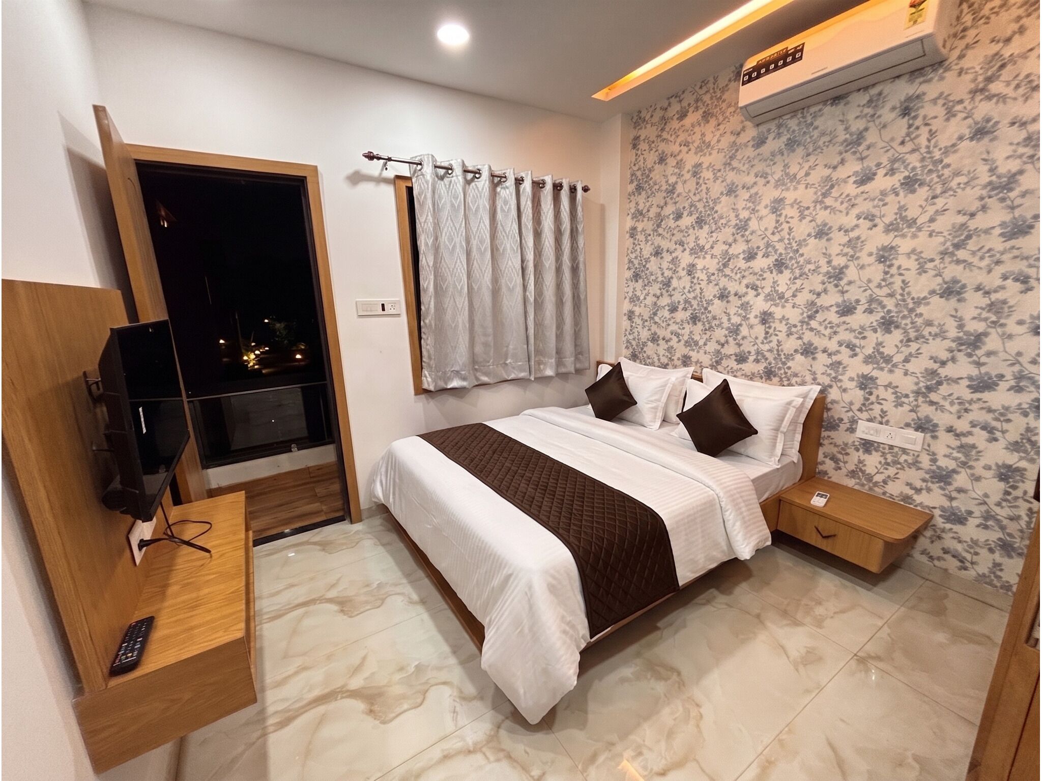 A hotel room with a bed, bedside tables, a TV, a window, and a wall with floral wallpaper.