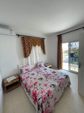 A bedroom with a bed, a nightstand, a window with curtains, and an air conditioning unit.