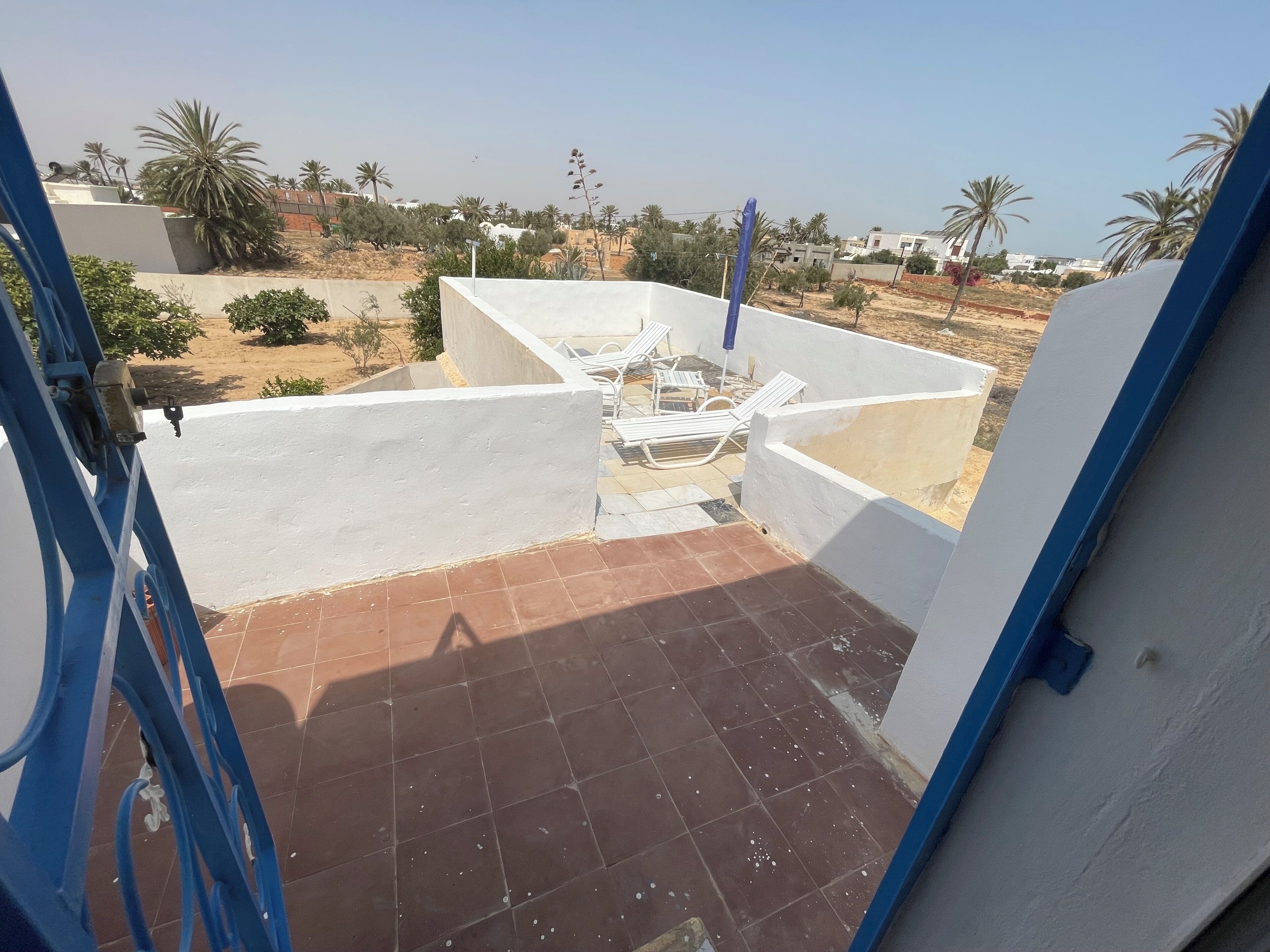 A rooftop terrace with a white railing, a blue gate, and a view of the surrounding area including trees and buildings.