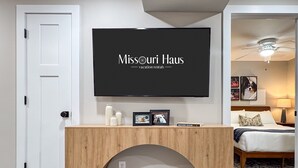 A hotel room with a wooden console table, a mounted TV displaying 'Missouri Haus vacation rentals,' a bed with a patterned pillow, and a nightstand with a lamp.