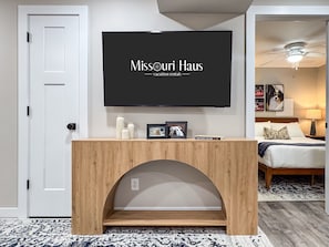 Ein Hotelzimmer mit einem Holzkonsolentisch, einem montierten Fernseher, der 'Missouri Haus Ferienwohnungen' anzeigt, einem Bett mit einem gemusterten Kopfkissen und einem Nachttisch mit einer Lampe.
