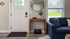 A modern living room with a white door, a round mirror, a wooden console table, a blue sofa, and a window with blinds.