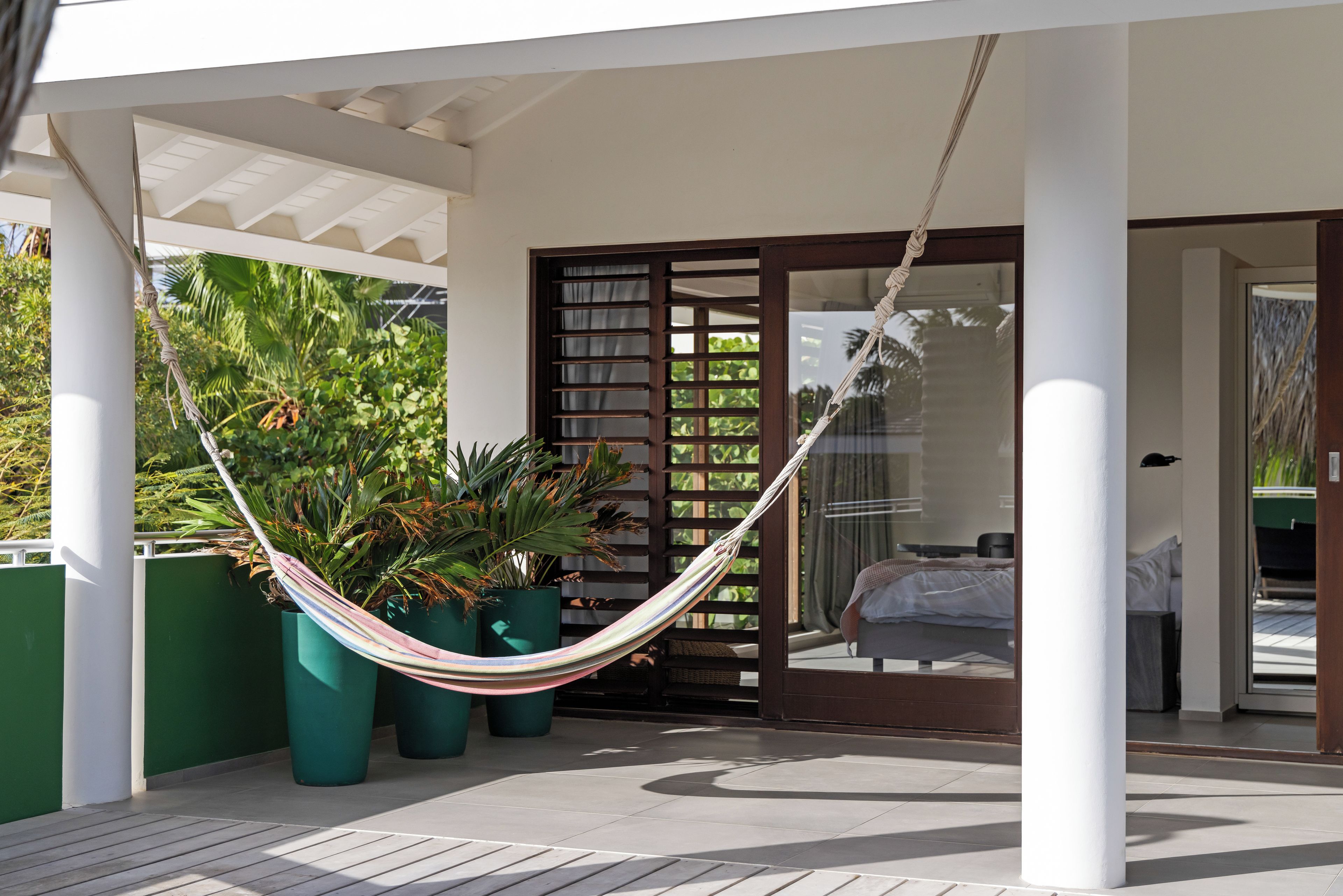 Una hamaca colgada entre dos columnas, una cama con cubrecama blanco y vista a un jardín exuberante a través de un ventanal.