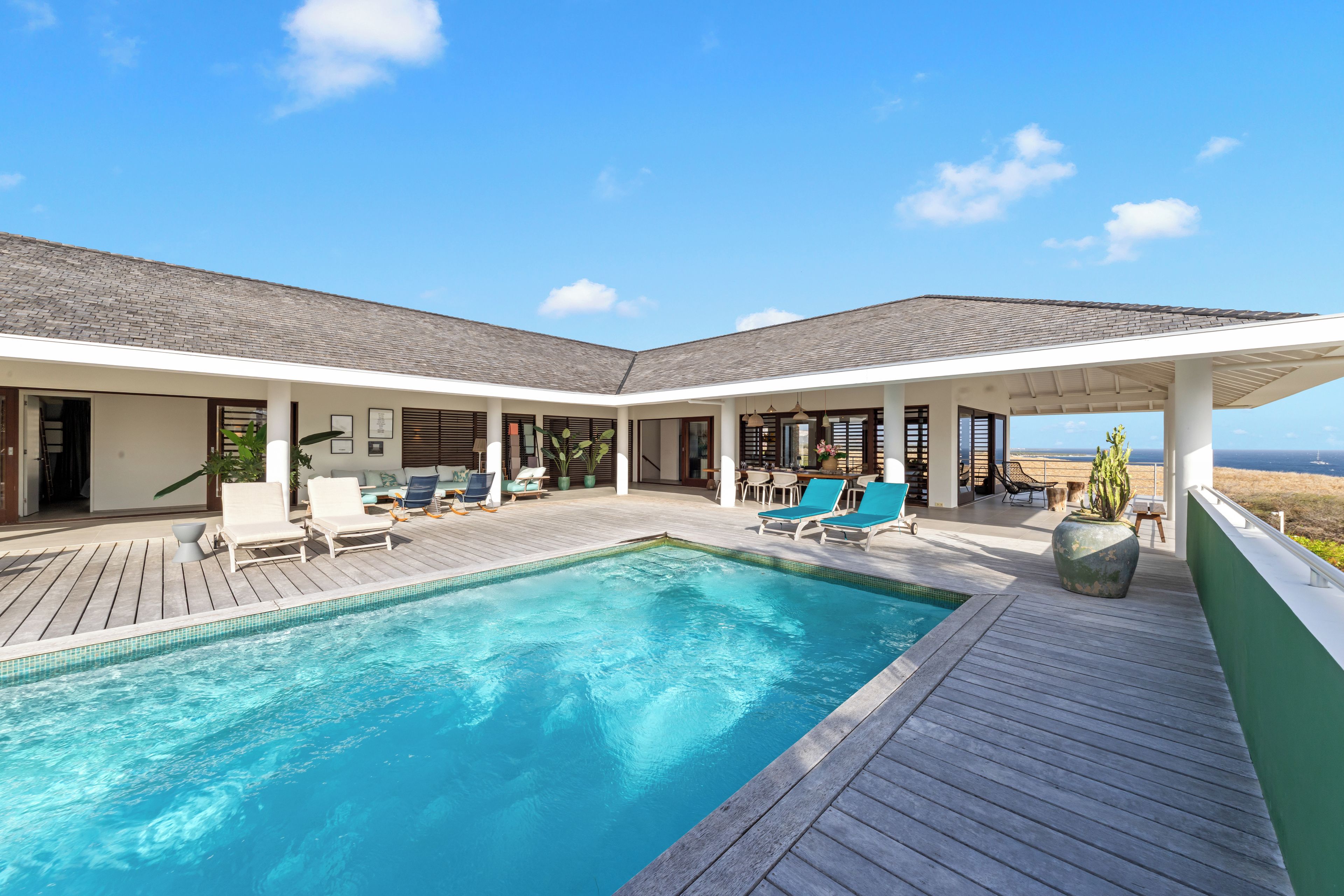 A poolside area with a clear view of the ocean, a covered seating area with lounge chairs, and a wooden deck.
