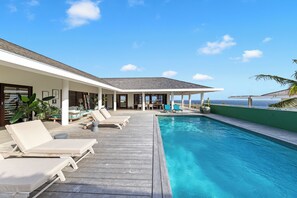 Un espace aménagé au bord de la piscine, avec des chaises longues, un coin salon abrité et une vue sur l’océan.