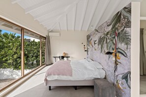 A bedroom with a large window, a bed with a white and pink blanket, a wall-mounted lamp, and a tropical-themed mural. - Villa Jan with swimming pool (Jan Thiel)