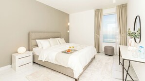 Ett modernt hotellrum med en stor säng, ett nattduksbord med en lampa, ett skrivbord med en stol och utsikt över havet.