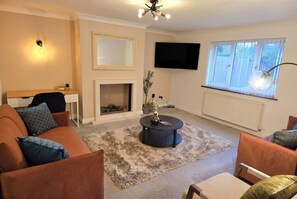 A living room with a sofa, armchairs, a coffee table, a fireplace, a TV, and a window with blinds.